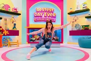 A girl (Lisette Xavier) crouched down with her arms outstretched. She is in front of a sign that says "Bestest Day Ever".
