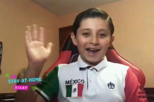 A headshot of a young boy waving. He is wearing a shirt that has a small mexico flag on it with the words Mexico.
