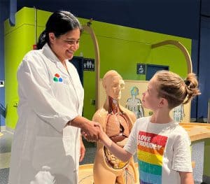 A woman in a lab coat is shaking hands with a young girl. Behind them is a plastic lifesize person with the internal organs visible.