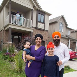 A young Sikh family. Wife holding a baby, husband and a young girl. They are outside a house, standing on the sidewalk. The house has a balcony and there is an older man and woman on the balcony.
