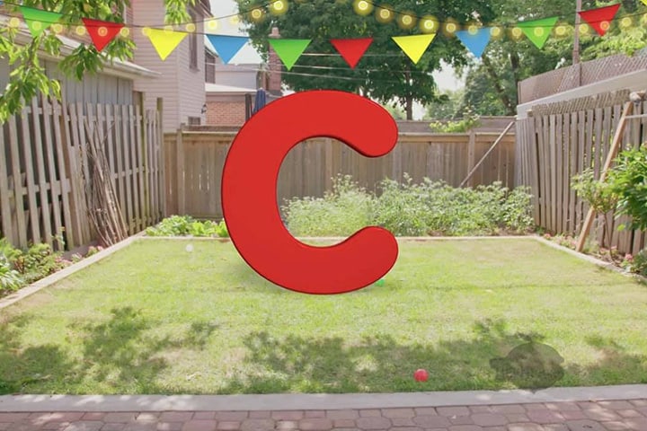 A fenced-in backyard with green grass. There is a string of coloured flags stretched across the yard. There is also a big letter "C" in the middle of the image.