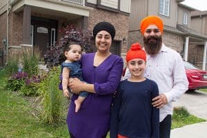 A young Sikh family. Wife holding a baby, husband and a young girl. They are outside a house, standing on the sidewalk. The house has a balcony and there is an older man and woman on the balcony.