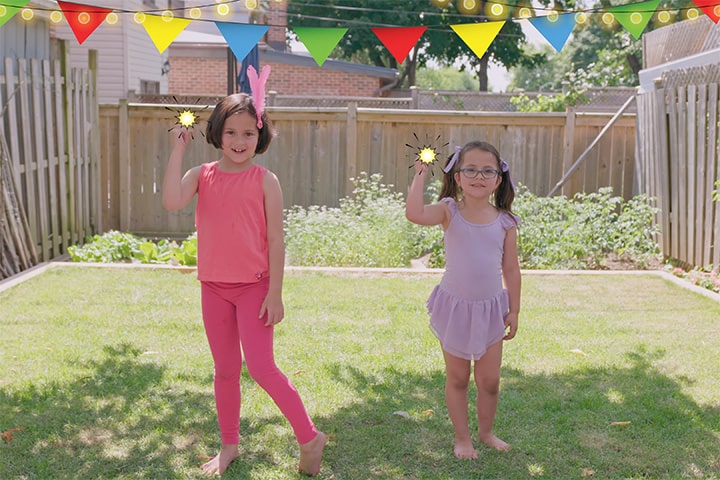 Two girls standing in a fenced in backyard. There is a bunch of colourful flags strung across the yard. The girls are each holding a glowing ball in their right hand.