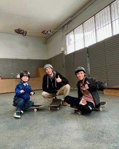 Tai Young sitting on a skateboard, with a skateboard instructor and a young boy, also on a skateboard. They are all giving the thumbs up sign. This is an episode of "Dream It Believe It"