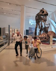 Tia Young (in a wheelchair) and a man and a young girl pretending to be dinosours. They are at a museum with dinosaur skeletons in the background. This is a scene in an episode of "Dream It To Be It".