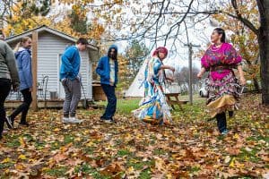 Two native americans in brightly coloured indian costumes dancing. Four teens are dancing with them. It is fall, with lots of leaves on the ground.